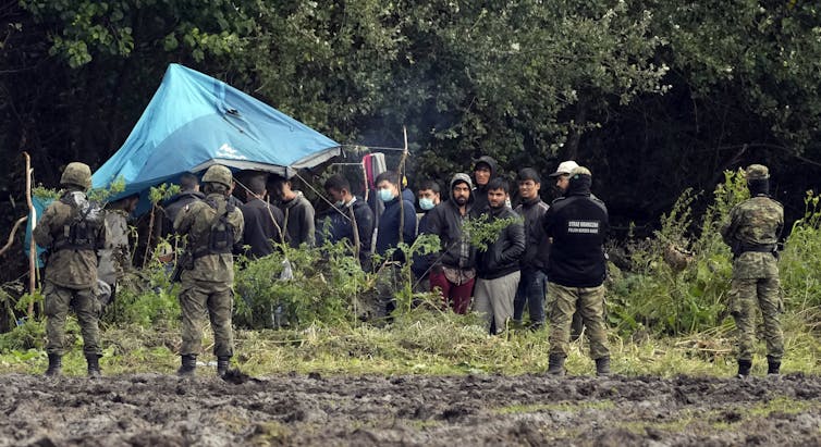 Men, some wearing masks, stand together and stare at armed soldiers in camouflage uniforms.