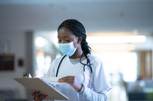 Young woman wearing face mask and scrubs looks at papers on a clipboard.