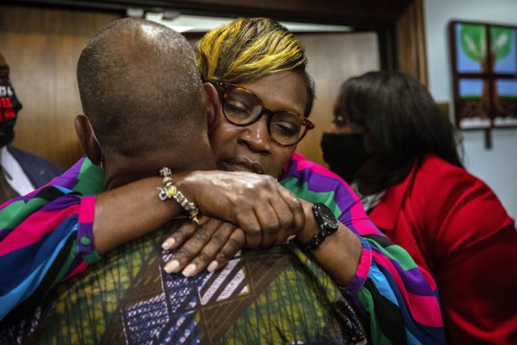 A Black woman wearing glasses embraces a man facing away from the camera.