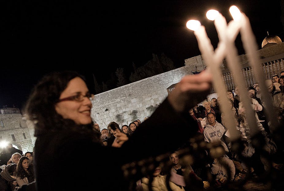A woman in a dark coat and glasses lights Hanukkah candles outside.