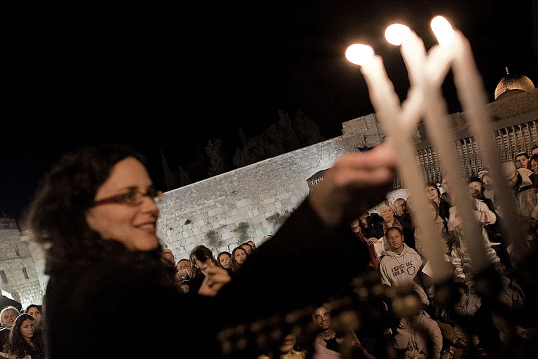 A woman in a dark coat and glasses lights Hanukkah candles outside.