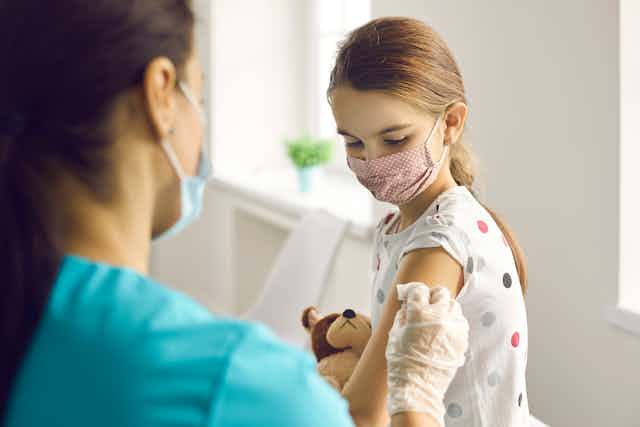 A nurse applies a bandaid after vaccianting a girl holding a teddy bear.