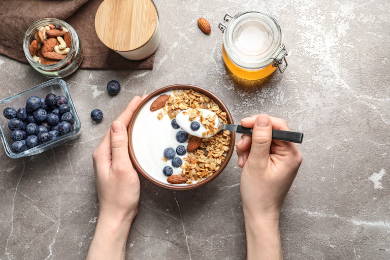 An overview of a bowl of yoghurt with blueberries and granola. A person's hands are holding the bowl and a spoonful.