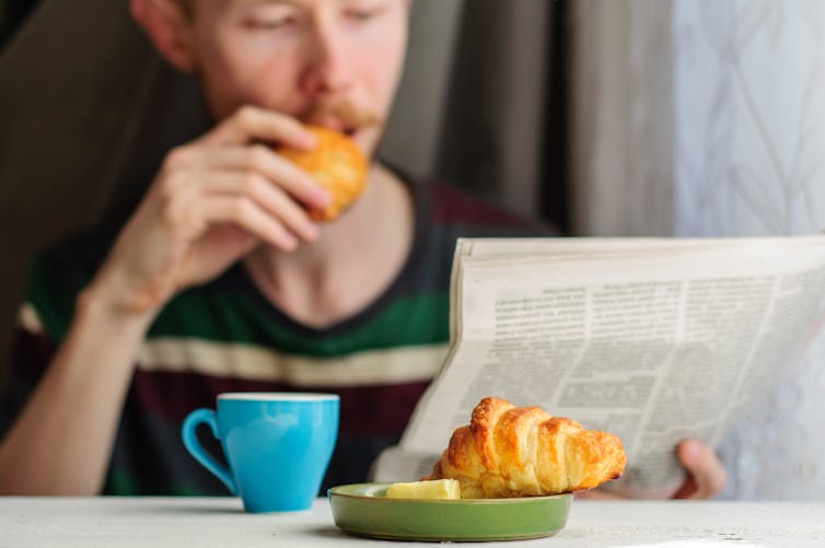 A young man in the blurred background eats a croissant while reading the newspaper. A cup of coffee and croissant are on the table in focus.