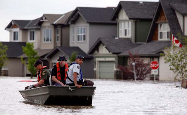 three men sit in a small metal boat as it moves past houses, a stop sign and a Canadian flag