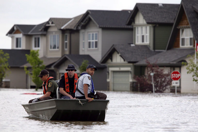 three men sit in a small metal boat as it moves past houses, a stop sign and a Canadian flag