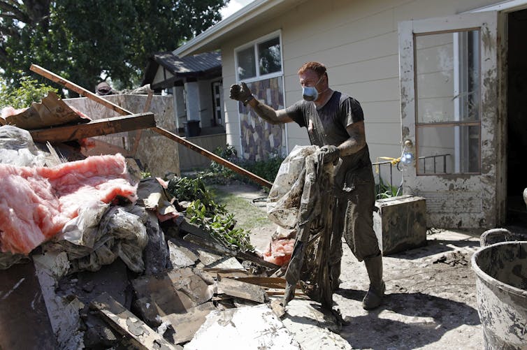 A man wearing a mask and work gloves throws muddy debris into a pile next to a house.
