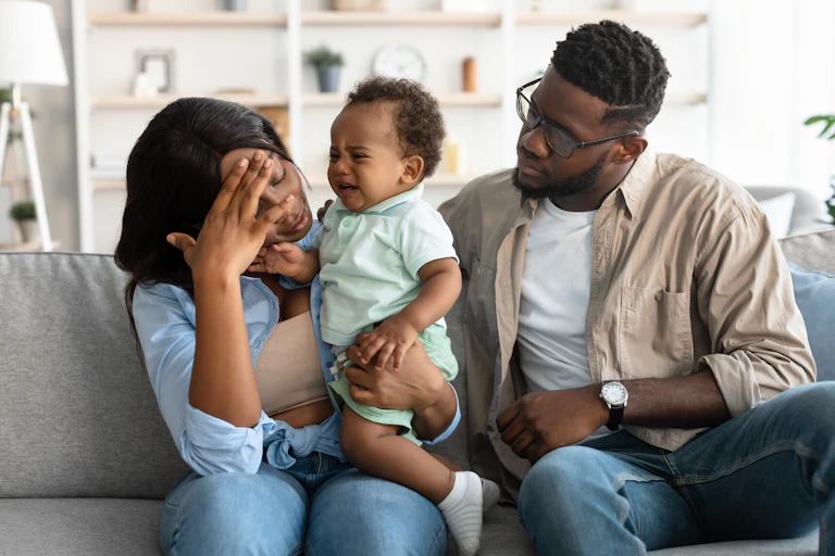 A frustrated mum holding a crying baby, while her partner sits nearby trying to comfort her.
