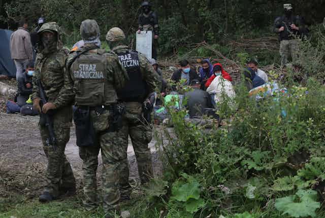 Armed Polish border guards standing beside a group of refugees sitting in a forest