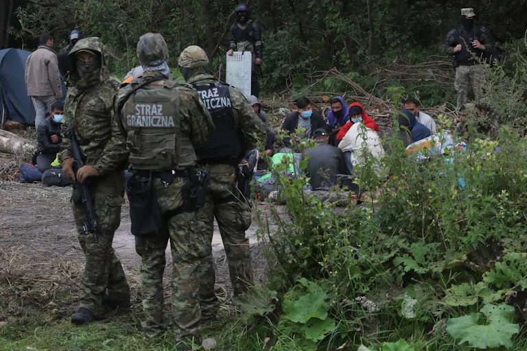 Armed Polish border guards standing beside a group of refugees sitting in a forest