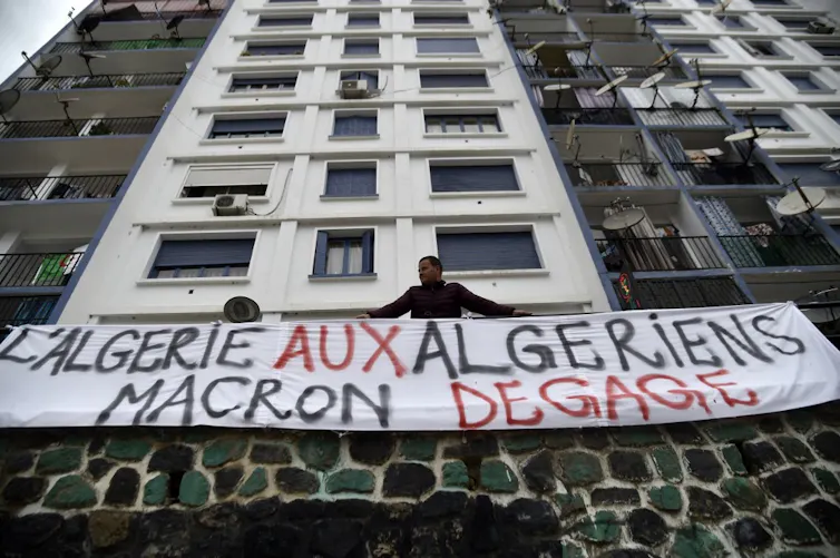 Un homme en bas d'un immeuble est appuyé contre une rambarbe sur laquelle est étendue une bannière blanche où il est écrit : L'Algérie aux algériens Macron dégage.