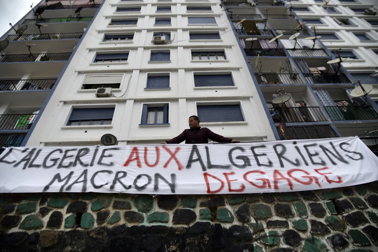 Un homme en bas d'un immeuble est appuyé contre une rambarbe sur laquelle est étendue une bannière blanche où il est écrit : "L'Algérie aux algériens Macron dégage."