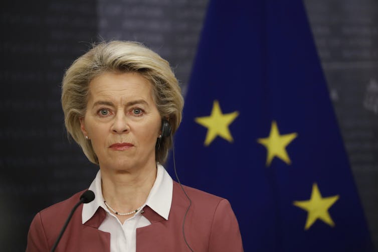 Ursula von der Leyen with a serious expression and an EU flag in the background