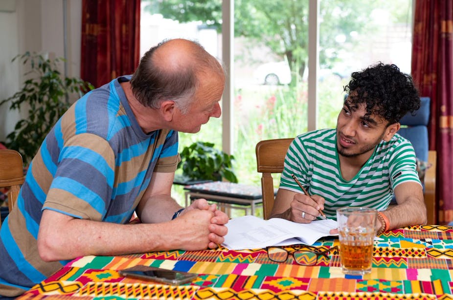 An older Dutch man and a young Syrian man sitting at a table and speaking to each other while the young man writes on a piece of paper