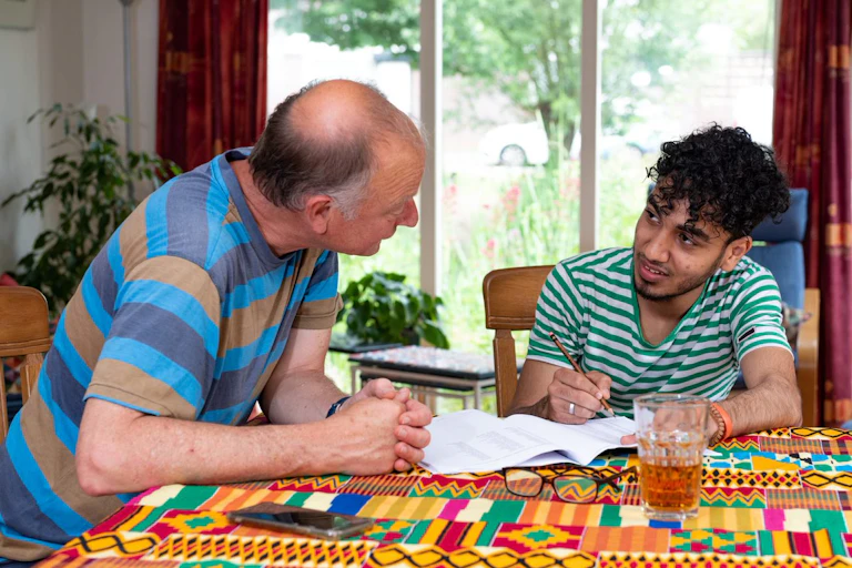 An older Dutch man and a young Syrian man sitting at a table and speaking to each other while the young man writes on a piece of paper