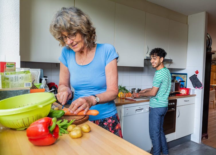 In the foreground, an older Dutch woman chops vegetables and smiles, while in the background a young Syrian man smiles and looks over his shoulder at her.