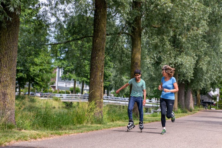 A young Syrian man rollerblades while his older woman host jogs alongside him on a tree lined path.