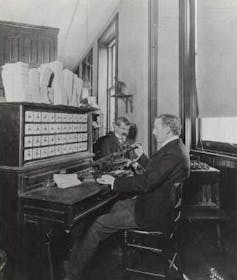 an old black and white photograph showing a man seated at a wooden desk-like machine looking at a bank of indicator dials