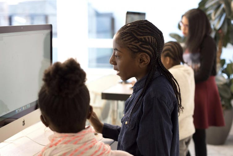 Black students are seen working at a computer with a Black teacher in the background.