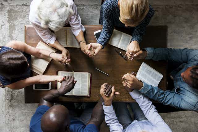 An overhead view of a group of six people praying with joined hands around a table with open Bibles in front of them.
