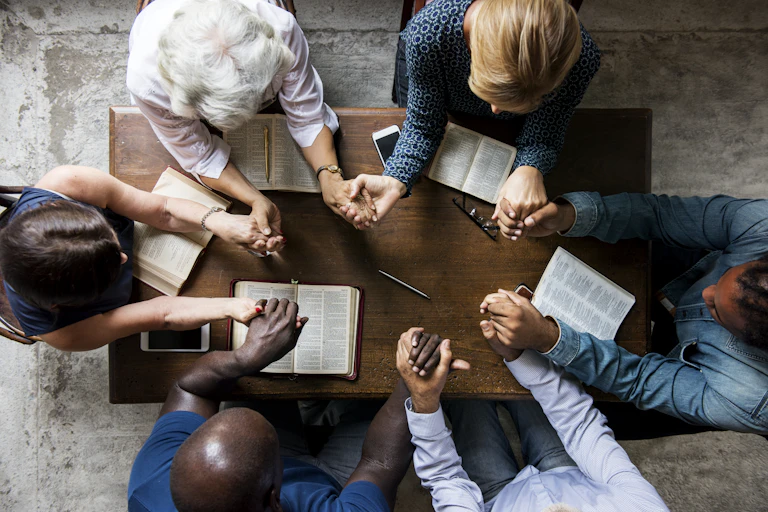 An overhead view of a group of six people praying with joined hands around a table with open Bibles in front of them.