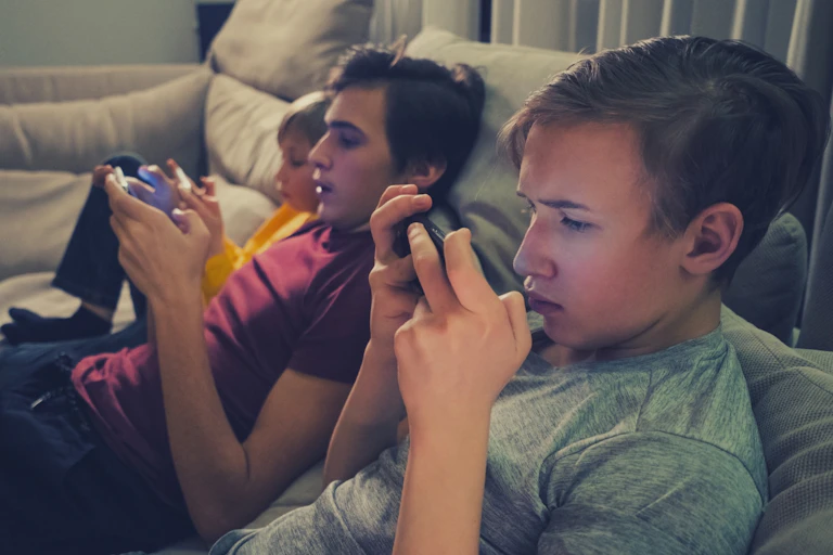 Three brothers of different pre-teen to teen ages are on the couch with devices.