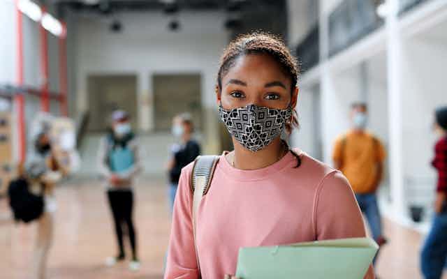A Black teen girl in a pink sweatshirt with a binder and and patterned face mask in a school corridor.