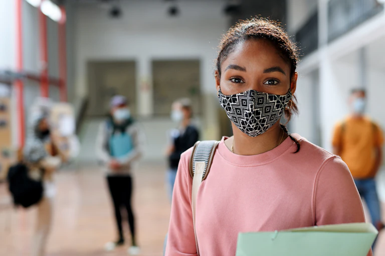 A Black teen girl in a pink sweatshirt with a binder and and patterned face mask in a school corridor.