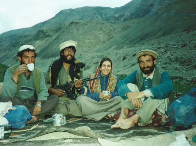 A woman in a headscarf and three men in turbans sit on the floor, smiling with a mountain in the background
