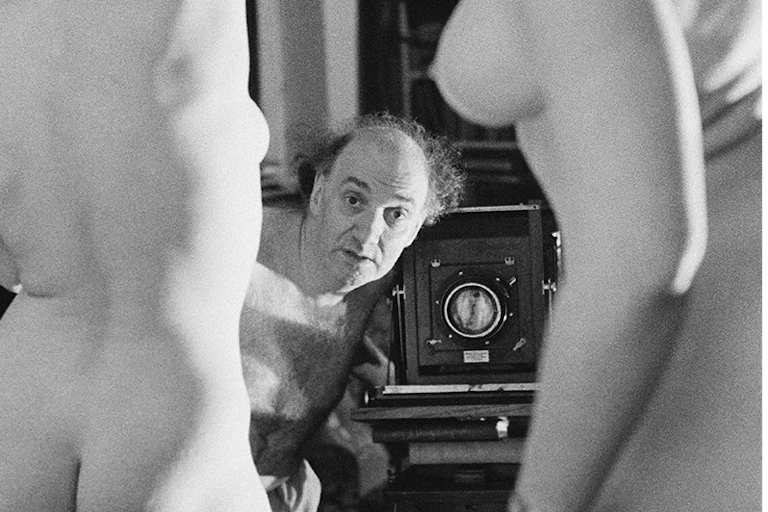A middle-aged man with a camera peers between the bodies of two young nude women