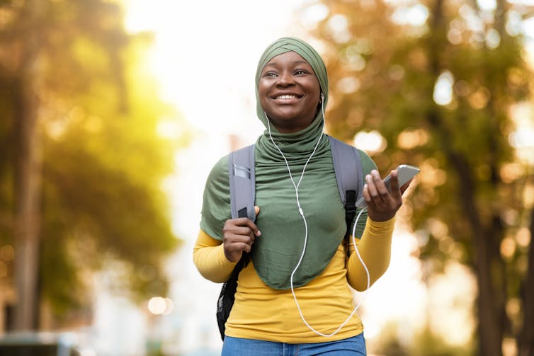 A girl wearing headphones and a hijab smiling while walking with a backpack.