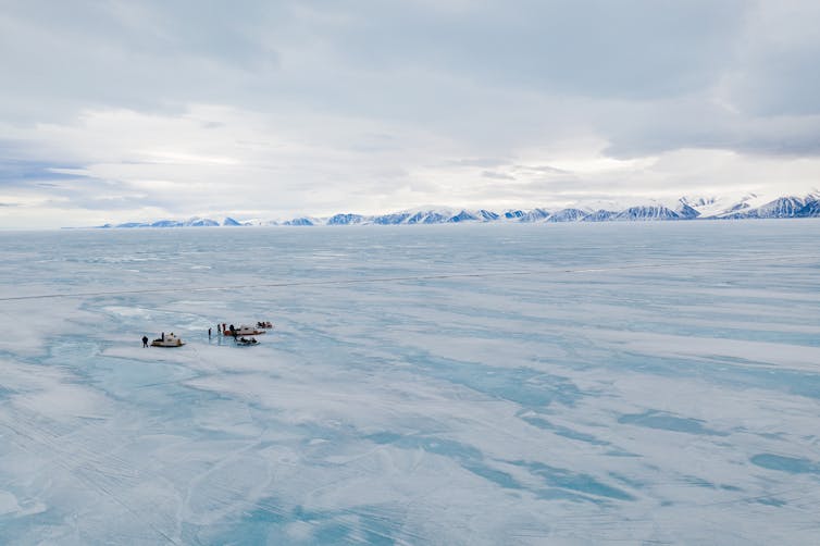 Aerial view of ice covered ocean with a line of mountain peaks in the distance. A small group of travellers are gathered near snow machines and wooden sleds.