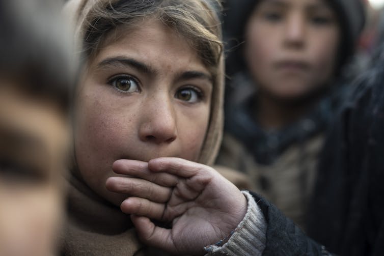 A girl with brown eyes and light brown hair looks at the camera, one hand against her mouth.