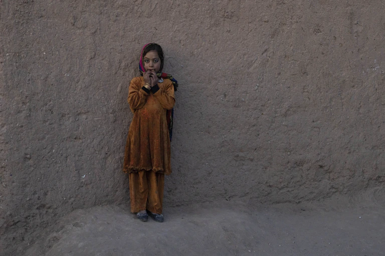 A girl in an orange outfit and headscarf looks at the camera with her hands on her mouth.