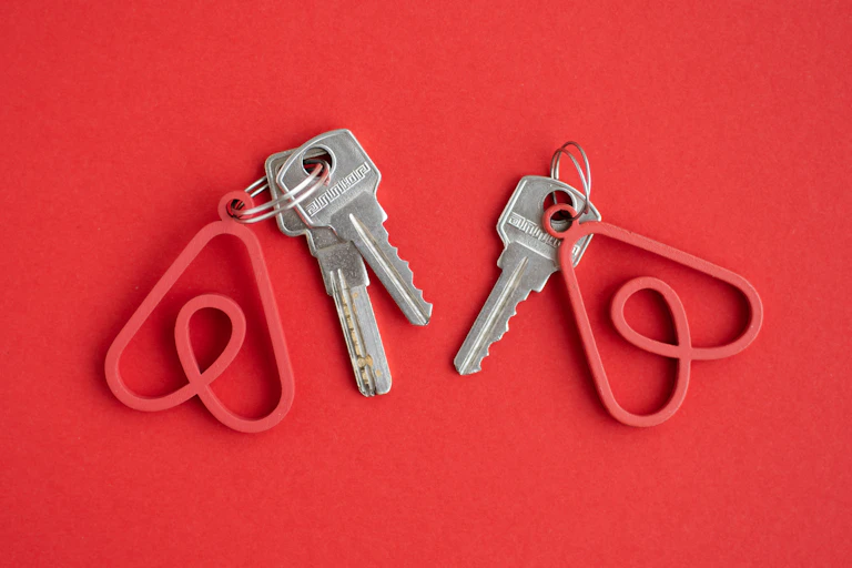 Two sets of keys with keychains in the shape of the Airbnb logo against a red background
