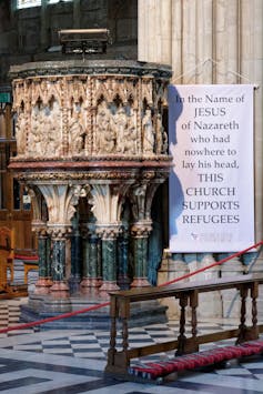 Ornate pulpit in the cathedral alongside which is a large banner hanging from a column in the name of Jesus of Nazareth proclaiming church support for refugees