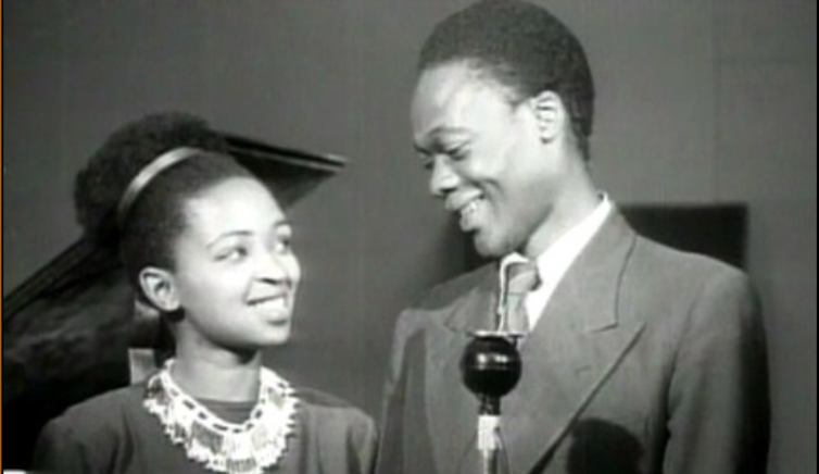 A glamorous man and woman smile as they share a microphone.