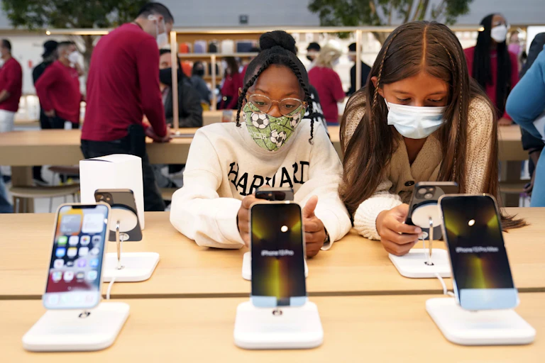 Two girls wearing masks hold and look at iPhones at an apple store
