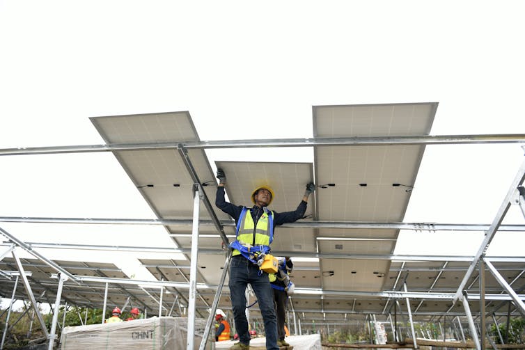 Um homem com um capacete de segurança fica sob três grandes painéis solares