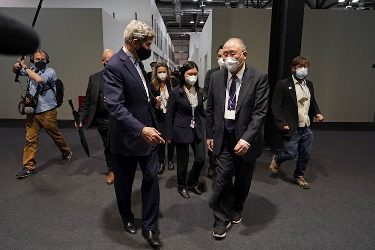 Negociadores americanos e chineses caminham por um corredor com repórteres atrás deles.