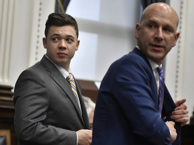 Two men in suits: on the left in grey is Rittenhouse and on the right in blue is his lawyer.