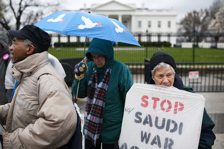 Sister Carol Gilbert, center, and Sister Ardeth Platte, right, standing with protesters in front of the White House holding a placard that says, 'Stop Saudi War.'