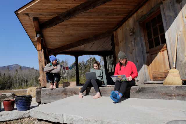 Three people sit on the porch of a rustic wooden home, two using laptop computers, with mountains in the background