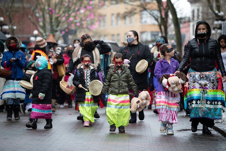 Girls and women in brightly colored skirts hold drums as they march.