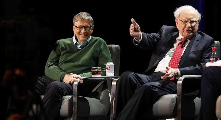 Bill Gates, in a green sweater, smiles while Warren Buffett speaks