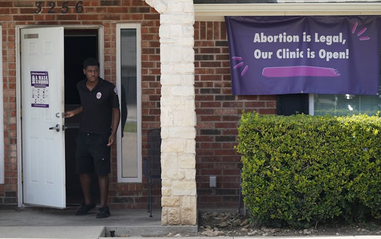 A security guard opens the door to a brick building with a green hedge in front in Fort Worth, Texas. A sign on the wall says abortion is legal our clinic is open