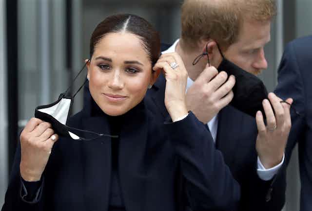 The Duchess of Sussex, Meghan Markle, taking off a COVID mask with her husband, Prince Harry, in the background also taking off a mask.