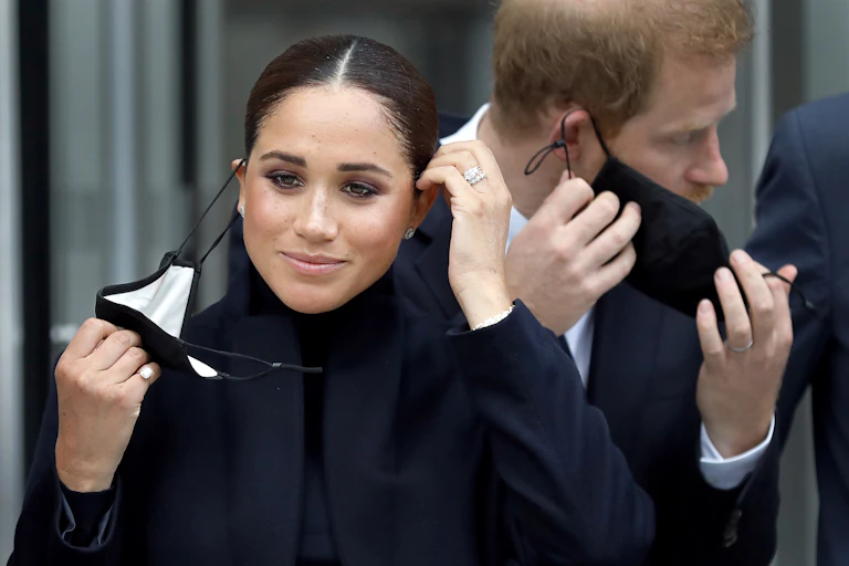 The Duchess of Sussex, Meghan Markle, taking off a COVID mask with her husband, Prince Harry, in the background also taking off a mask.