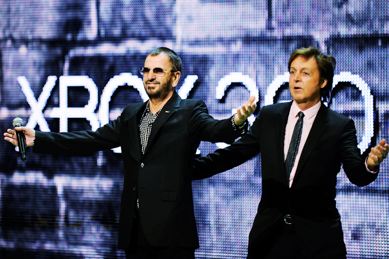 Two men in suits stand next to one another with their arms outstretched.