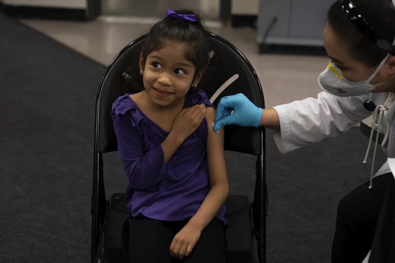 A six-year-old girl in a purple shirt and hair ribbon smiles as her arm is swabbed with alcohol